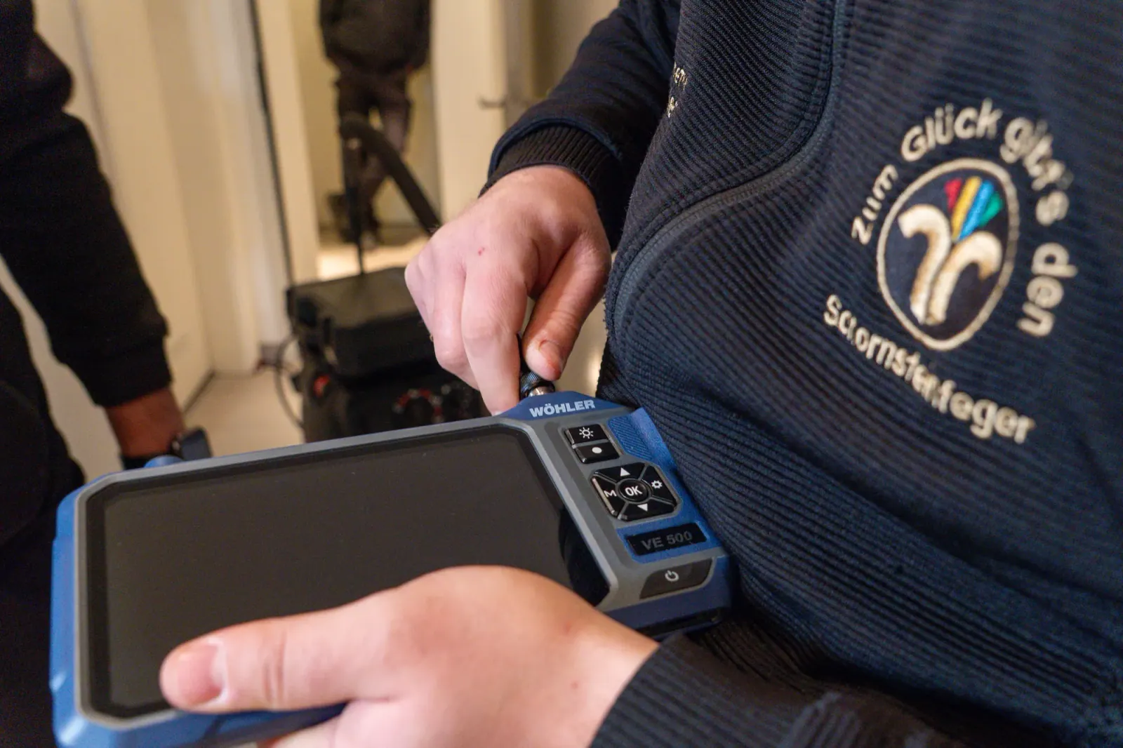 Hände halten ein Messgerät, Jacke mit Aufdruck „Zum Glück gibt's den Schornsteinfeger“